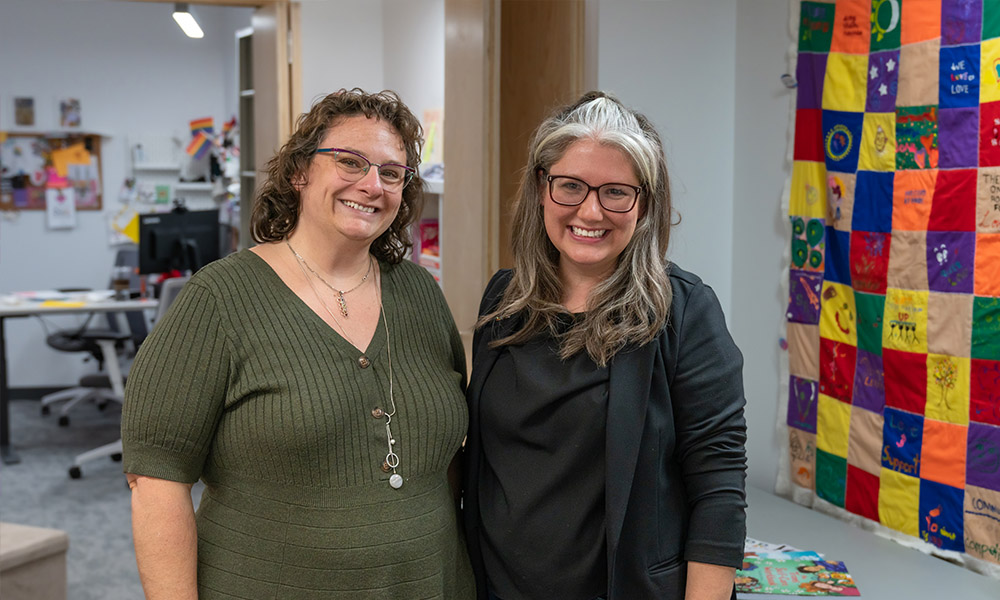 Jessica Scalzo and Kristie Benson stand alongside a colourful hanging quilt in their office space in the Centre for Sexual and Gender Diversity