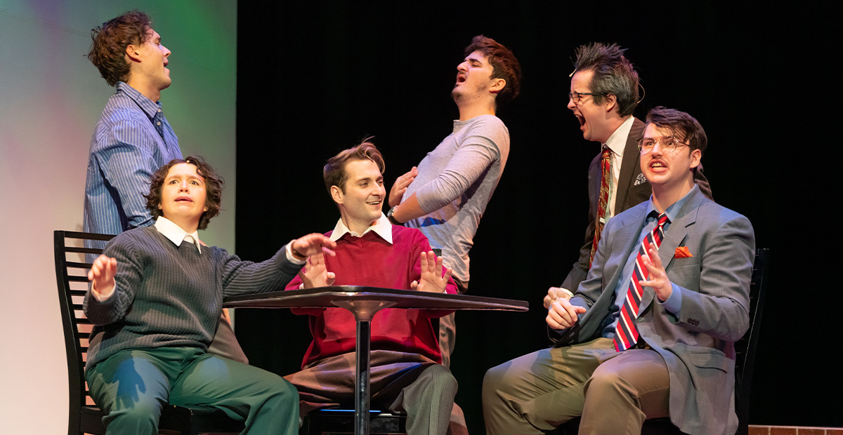 A group of men sit around a table singing during a production of the musical Company.