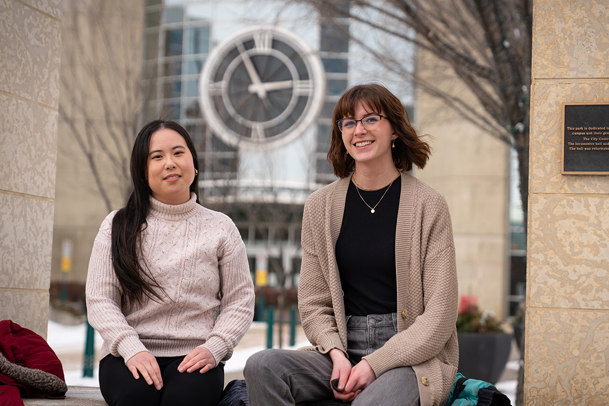 Shannon Lee and Grace Zimmel sit on a bench outside of MacEwan's clock tower entrance