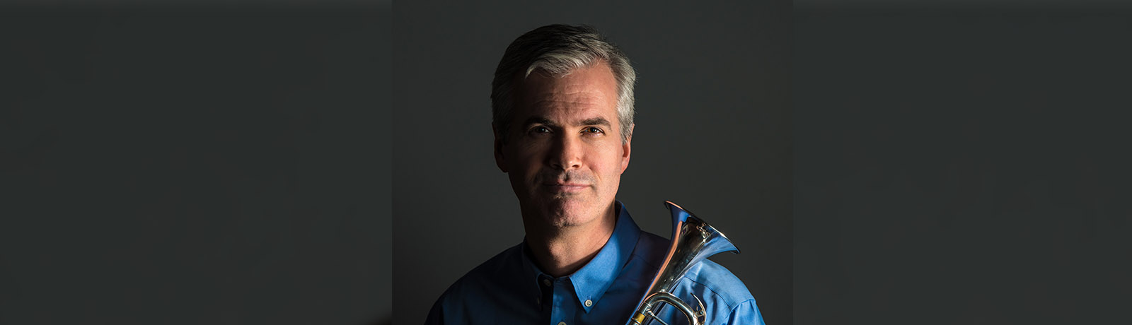 A close-up of Dean McNeill wearing a blue dress shirt and holding his trumpet