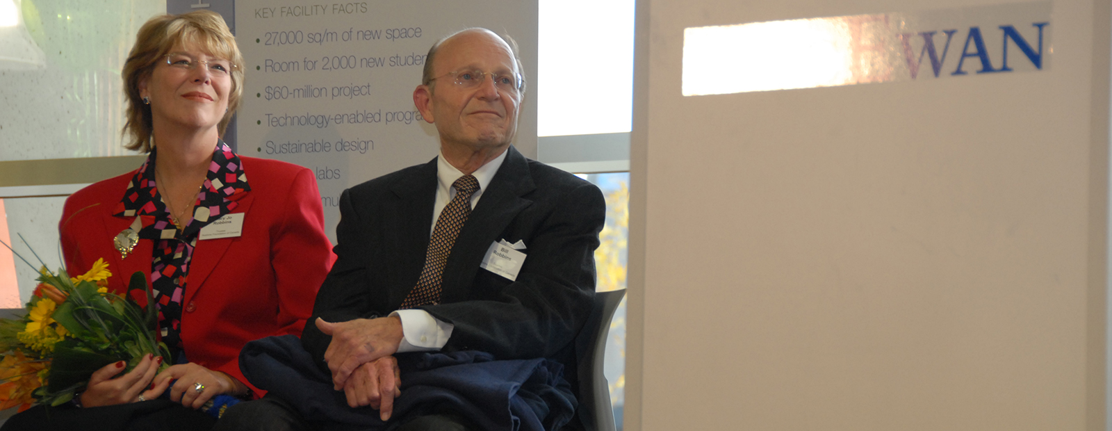 Bill and Mary Jo Robbins are seated in the Robbins Health Learning Centre. Mary Jo wears a red jacket and holds a bouquet of yellow flowers. Bill wears a dark blue suit. Both are looking to the right and smiling slightly.