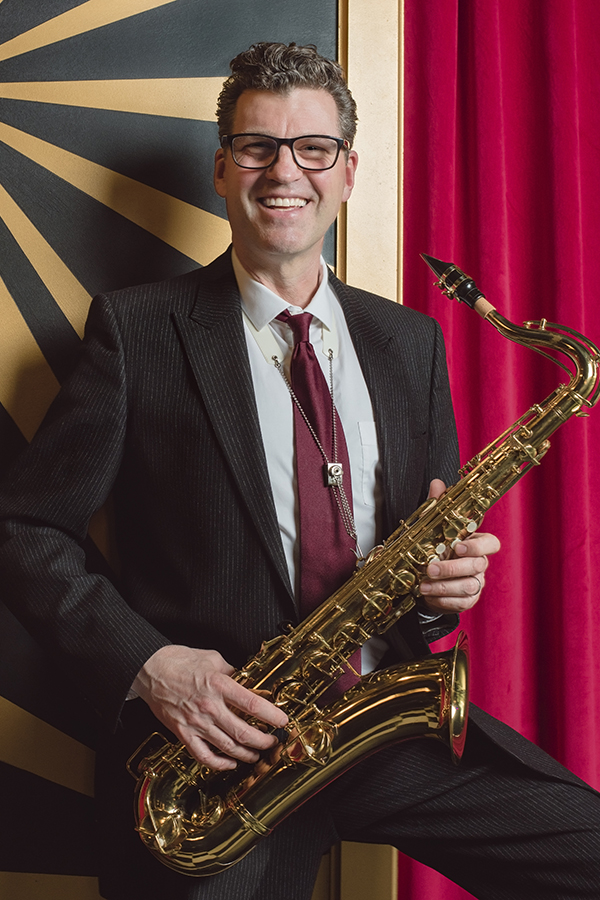 Dave Babcock is standing against a striped backdrop and holding a saxophone.