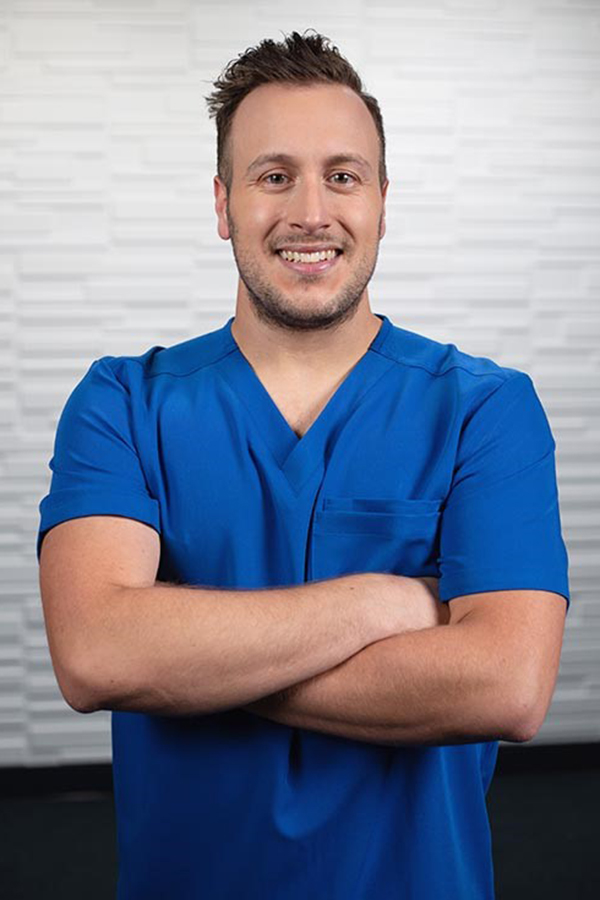 Brennan Belliveau is wearing blue scrubs and standing with his arms folded.