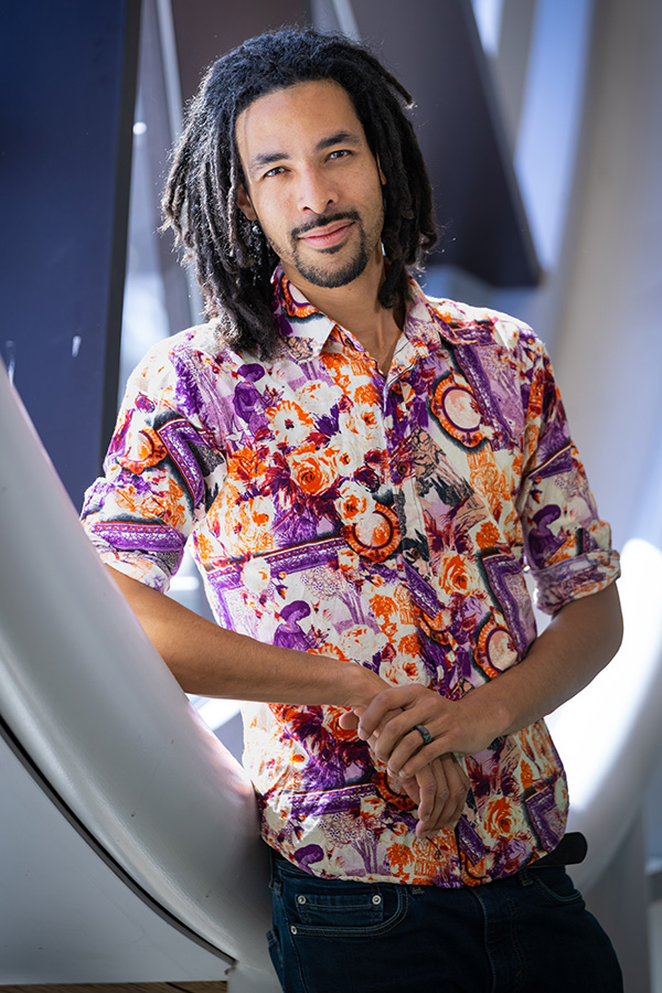 AJA Louden leans against a round window. He is wearing a brightly coloured floral shirt.