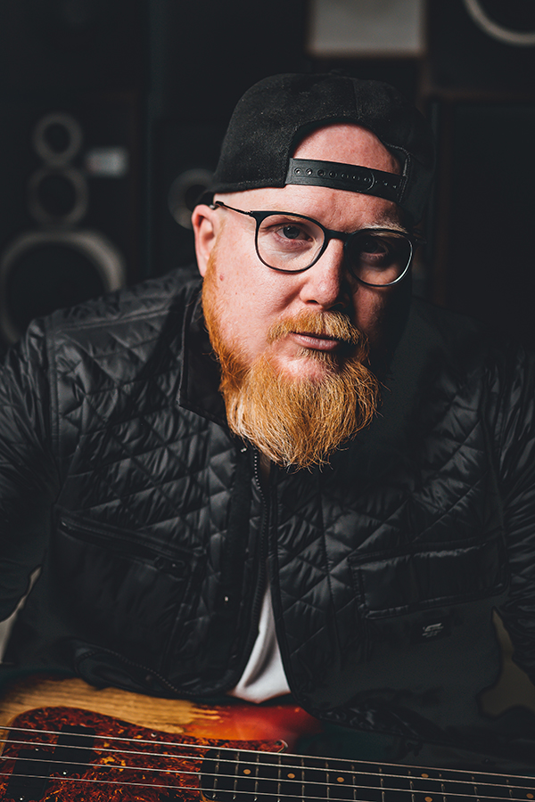 Travis Switzer wears a black jacket and a backwards black baseball cap. He is holding an electric guitar.