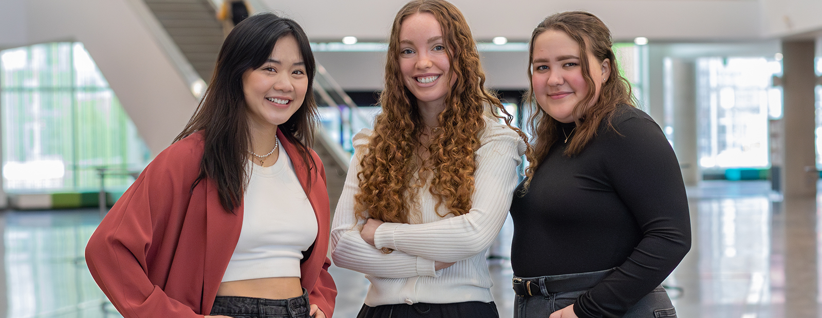 Three young women stand together in Allard Hall at MacEwan University. On the left is Mandy Mah, with long, straight, dark hair and bangs, and wearing a drapey red jacket.  In the centre is Eva Driessen, who has long red hair in ringlets and is wearing a white cardigan. On the right is Breanna Kitchen, whose brown hair is pulled into a half-ponytail. She is wearing a black turtleneck. All three women are smiling at the camera.