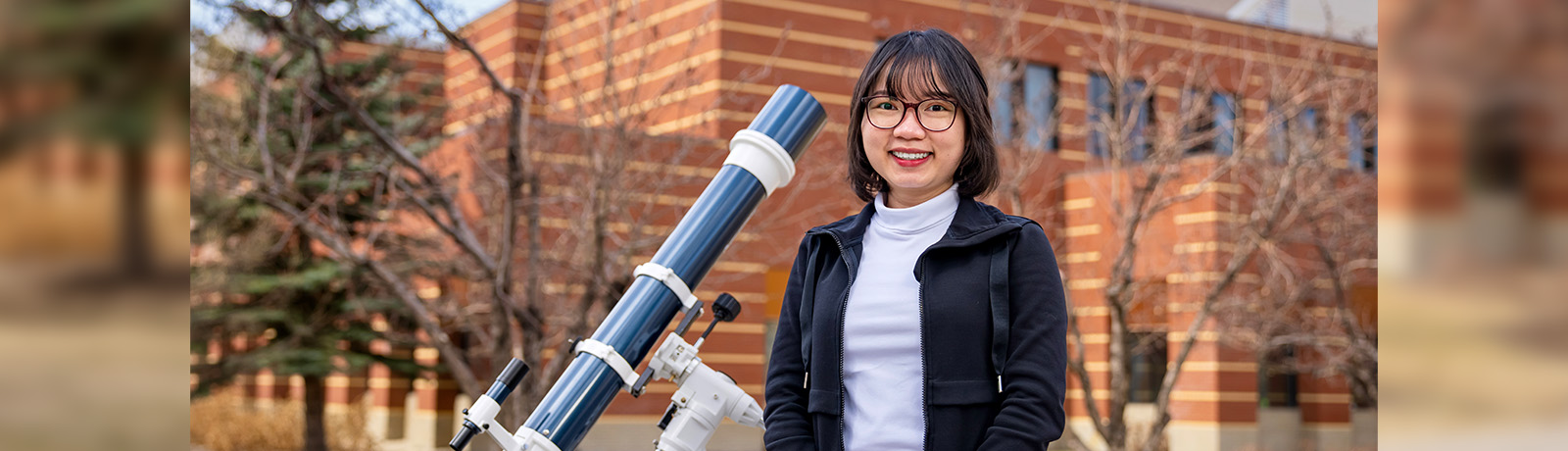 Vy Tran stands in front of Building 5 beside a large telescope