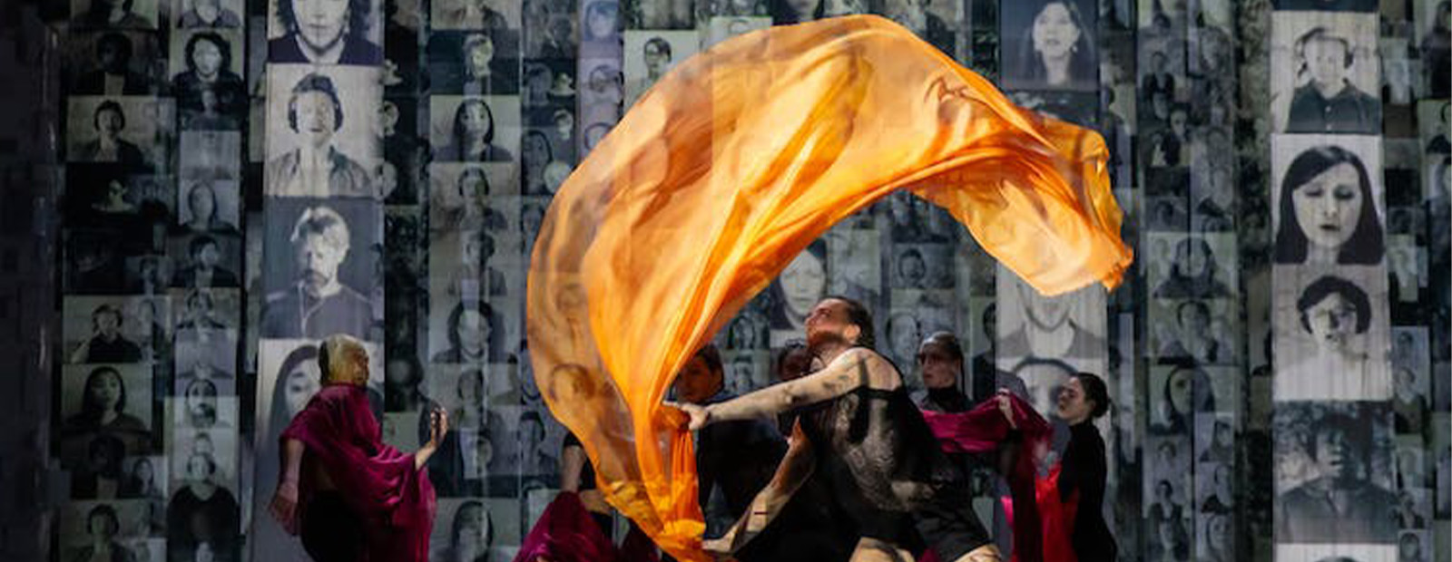 Performers use red and orange fabric to create a visual representation of fire while images of dozens of faces are projected onto the wall behind them.