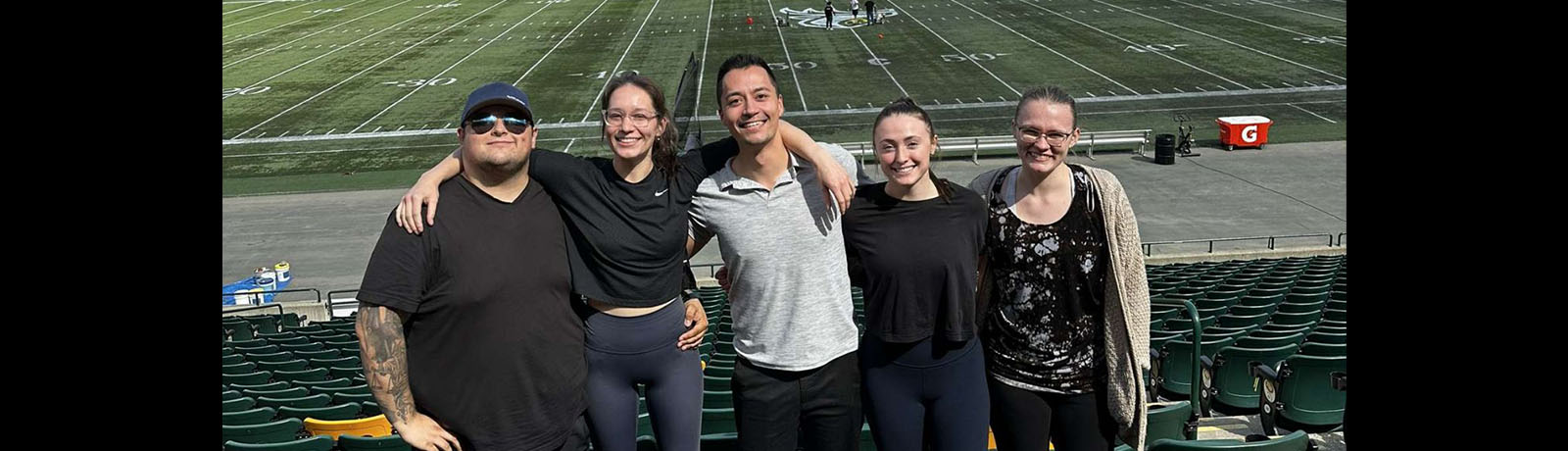 A group of five massage therapy students stand in among the seats in Commonwealth Stadium with the playing field in the background