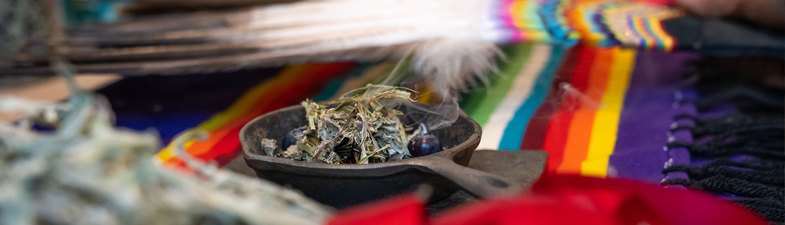 A cast iron pan holding sweetgrass is surrounded by vibrant cloth and eagle feathers