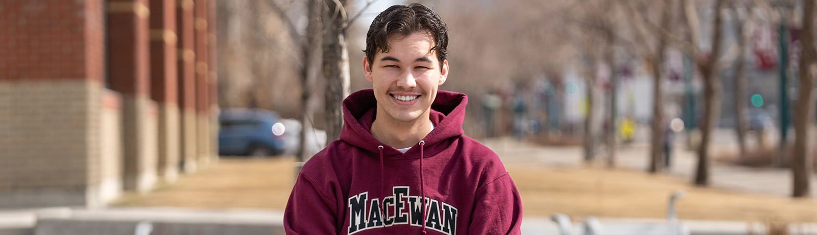 Conor McGhee sits on a bench outside of Building 7 wearing a MacEwan Police Studies hoodie