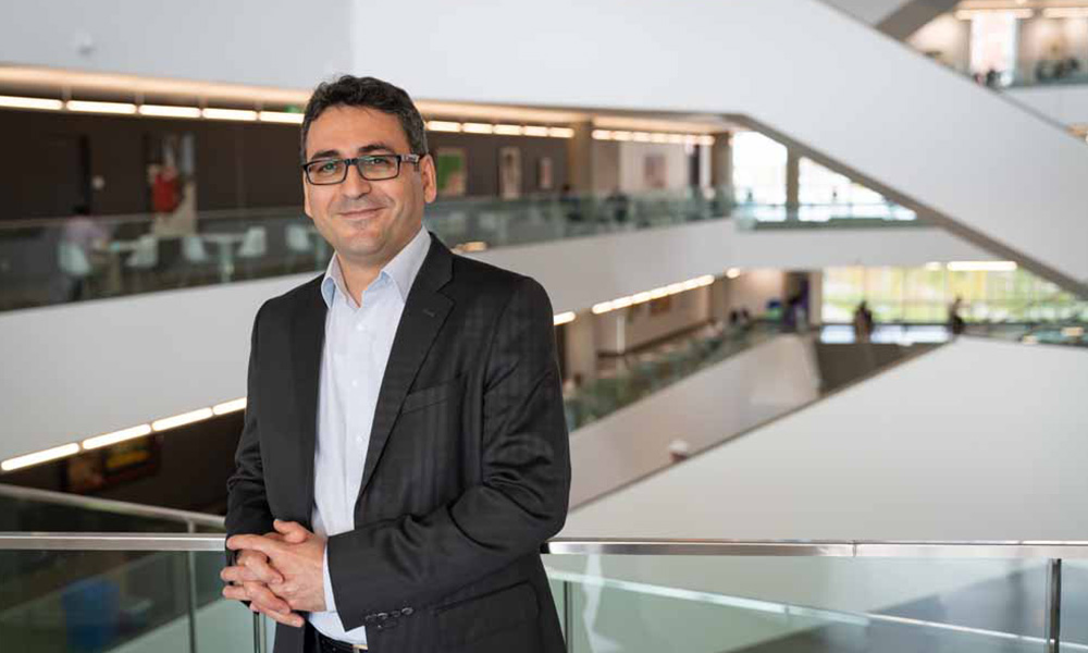 Dr. Chapardar wears a light blue shirt under a dark grey suit jacket as he leans on a railing in Allard Hall