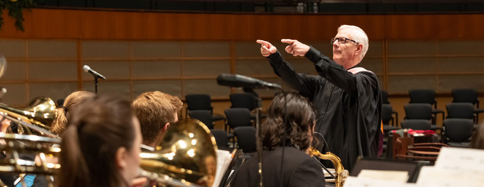 Ray Baril holds up his arms as he conducts the Big Band on stage at convocation, with the band in the foreground of the image facing Baril