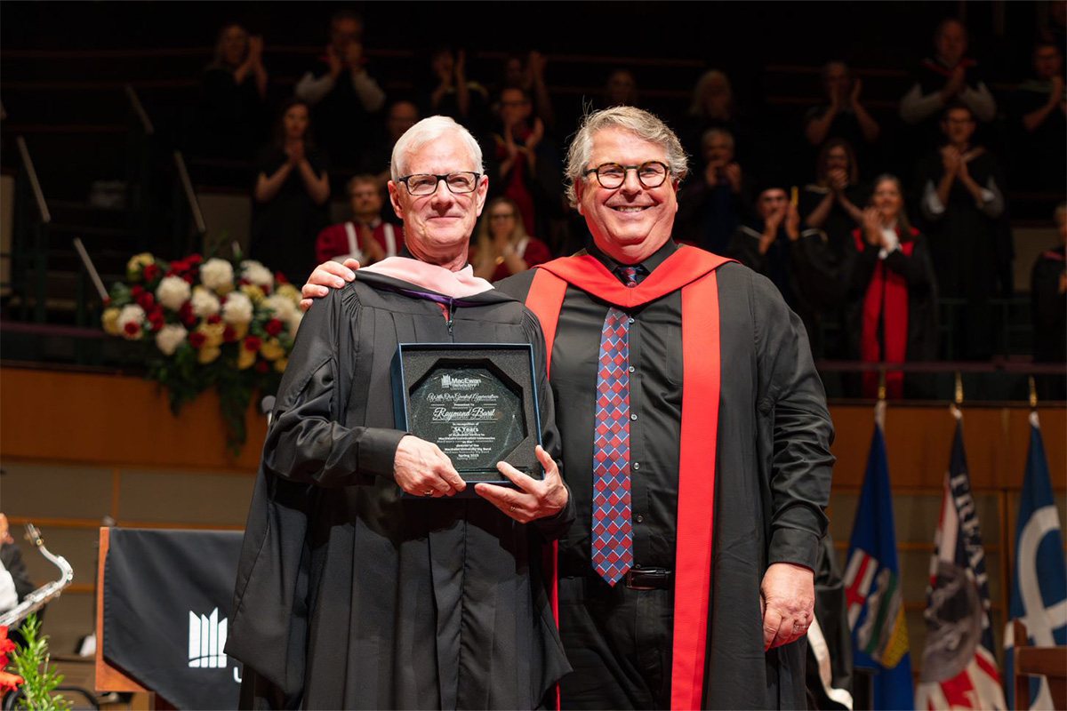 Ray Baril and Dr. Allan Gilliland stand together on stage at convocation, dressed in ceremonial robes. Ray holds an engraved glass plaque