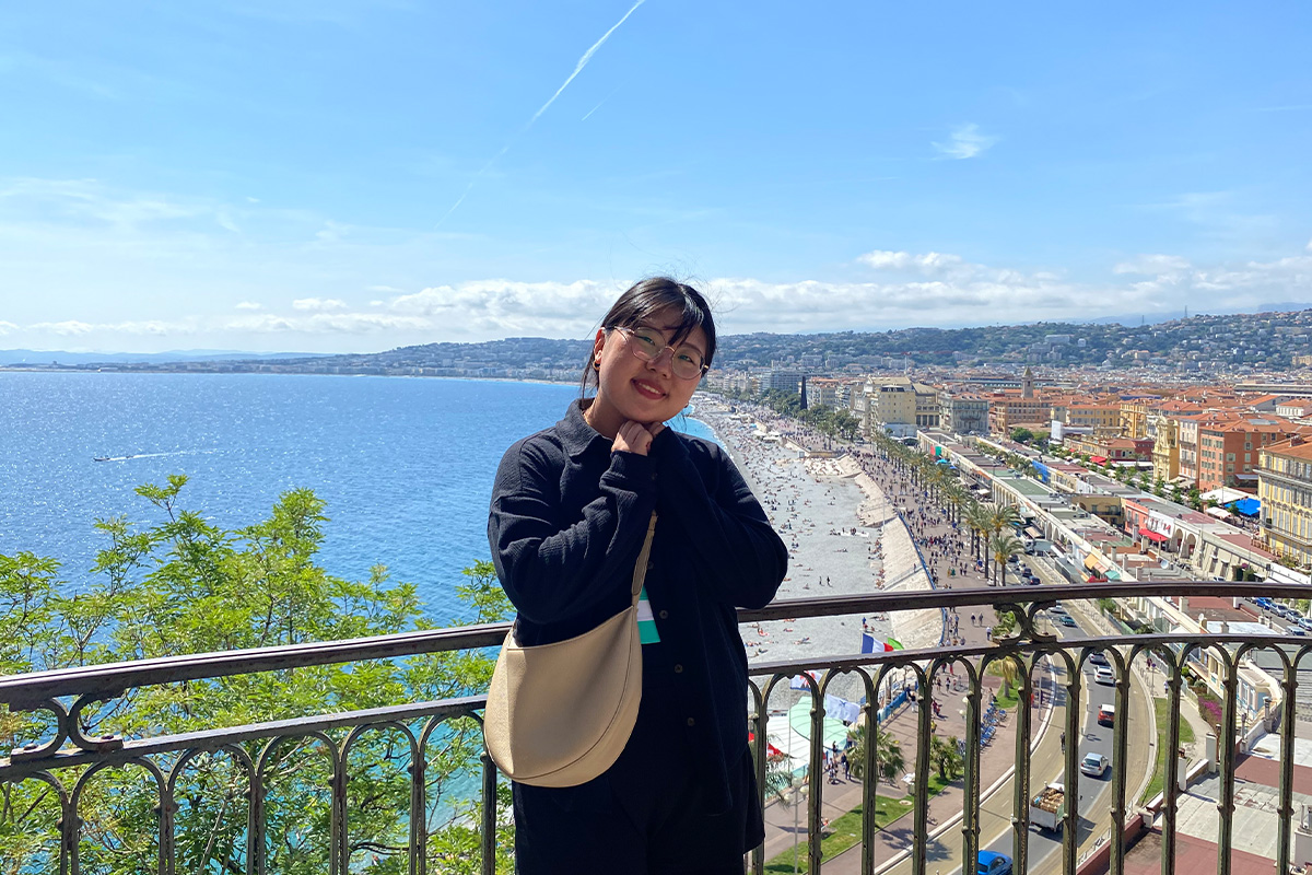 A woman with black hair stands on a balcony overlooking the edge of a city and a sea, with her hands folded beneath her chin