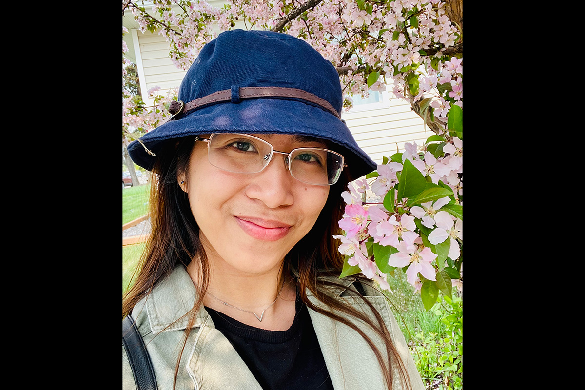 A woman in a blue bucket hat wearing glasses stands in front of a tree with blooming cherry blossoms