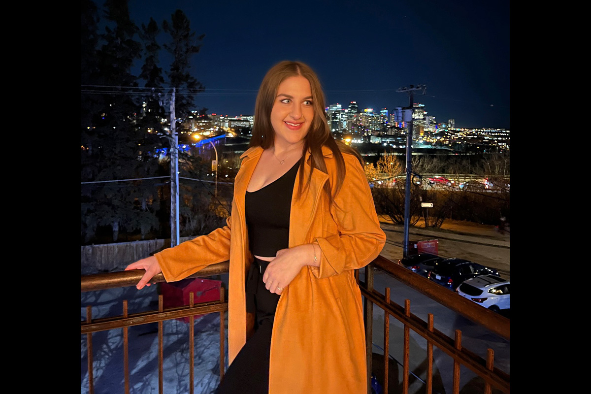 A woman in an orange jacket stands on a balcony overlooking the river valley and downtown at night