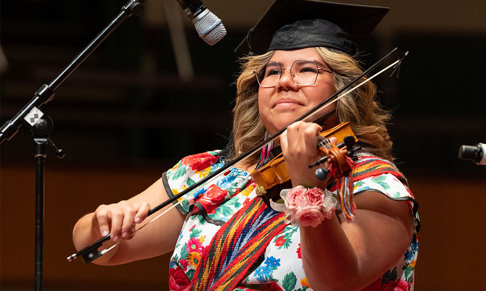 Brianna smiles while playing her fiddle on stage wearing her graduation cap