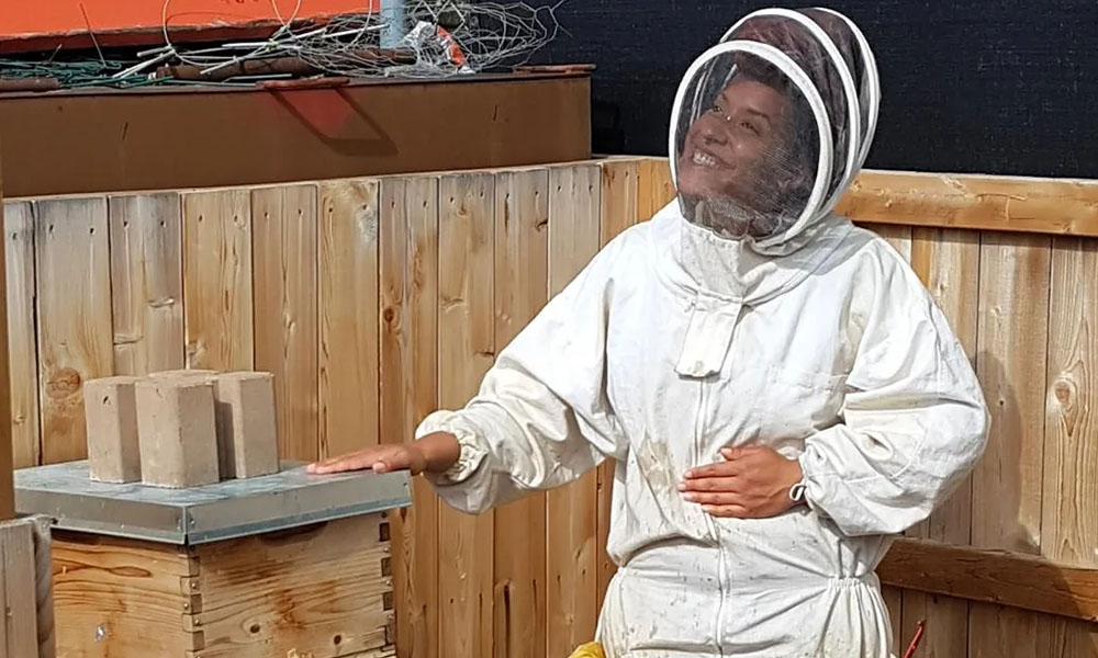 Amelia dressed in her bee suit, smiling with one hand resting on a hive