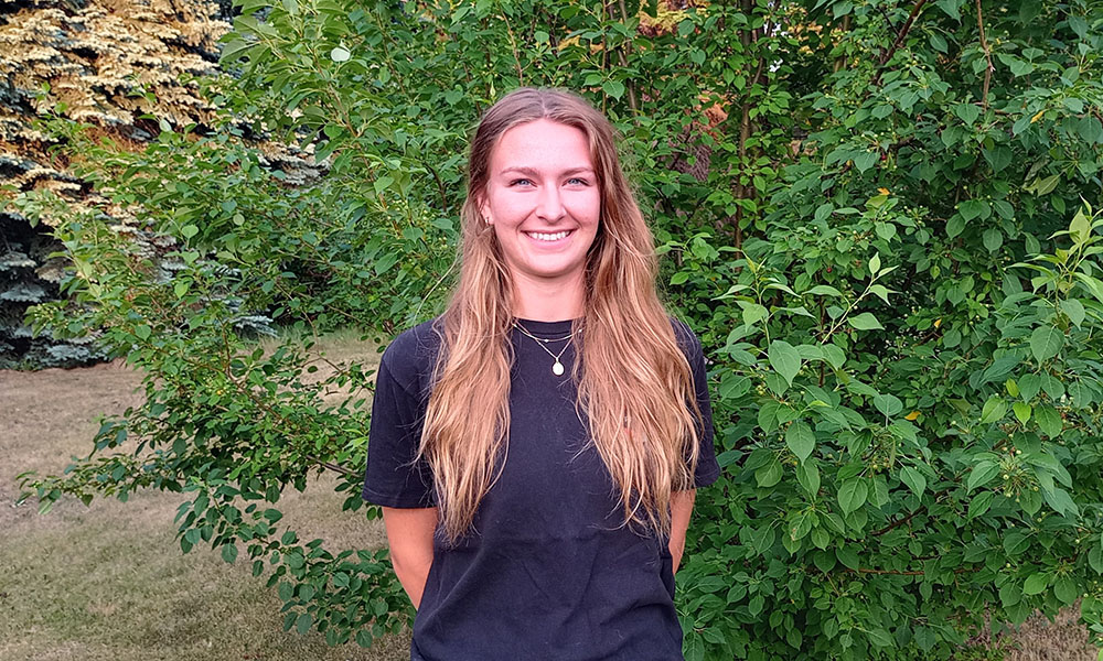 Shannon smiles while standing in front of a backdrop of green shrubbery