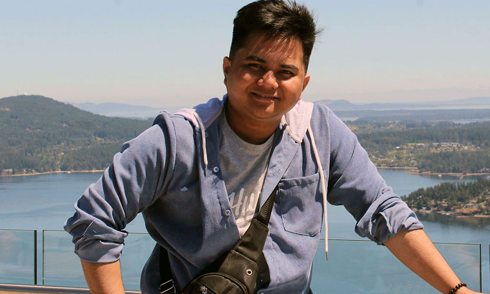 Christian stands on a walkway in front of water and rolling, tree-covered hills