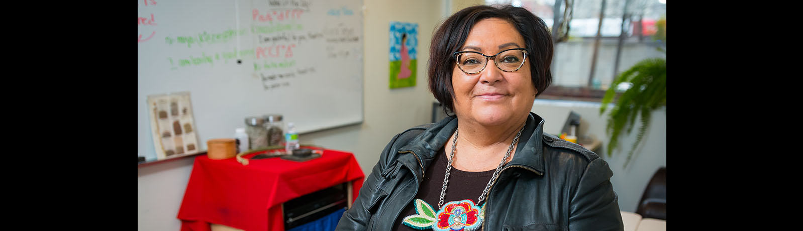 Roxanne Tootoosis stands in the former home of kihew waciston in Buiding 7 wearing a black jacket and beaded flower necklace in bright colours