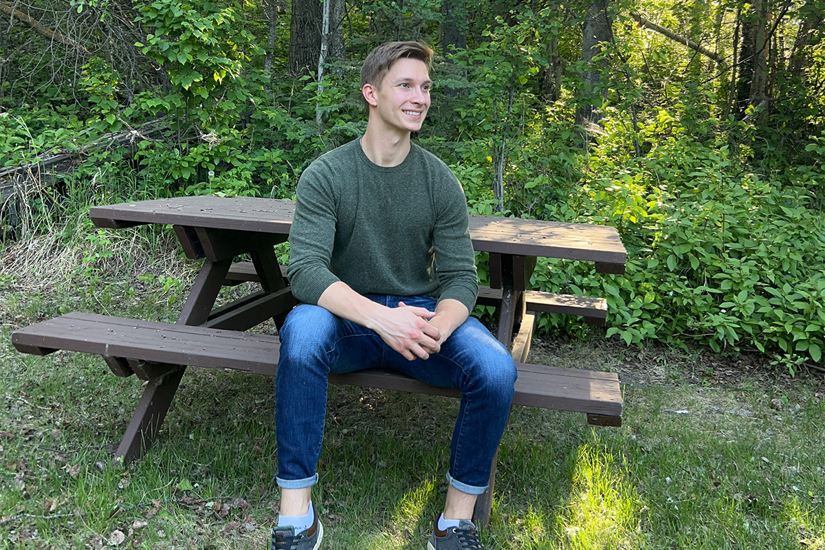 A man with short light hair sits on a wooden picnic table in front of vibrant green trees