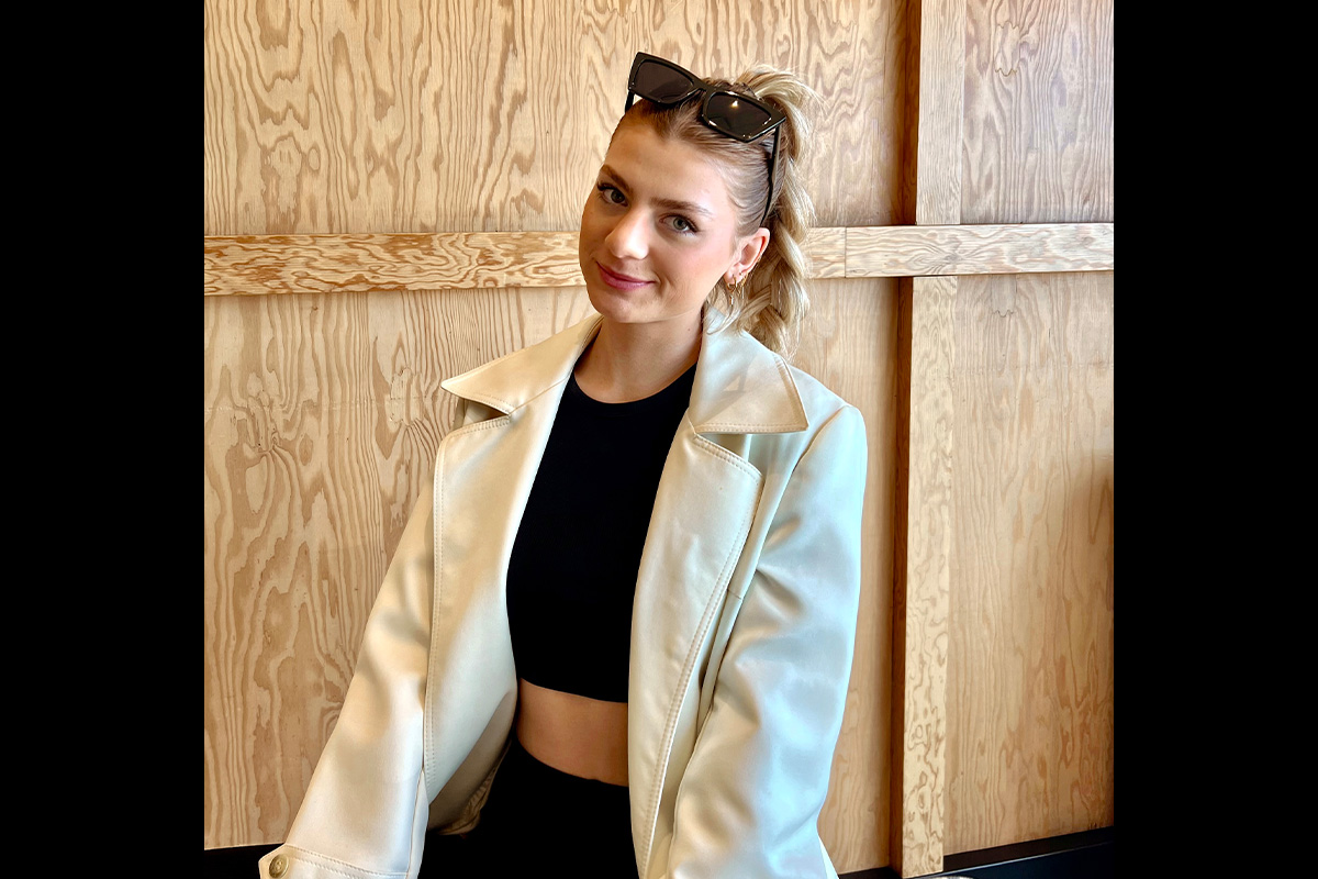 A woman with a blonde braid wears a light blazer and stands in front of a wooden wall