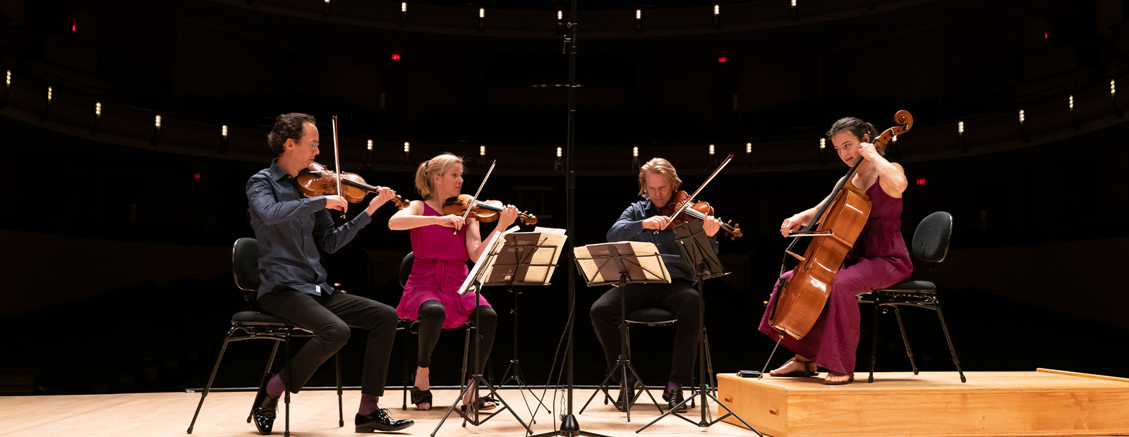 Two men and two woman sit on a stage, with one woman playing a cello while the other three musicians play violins.