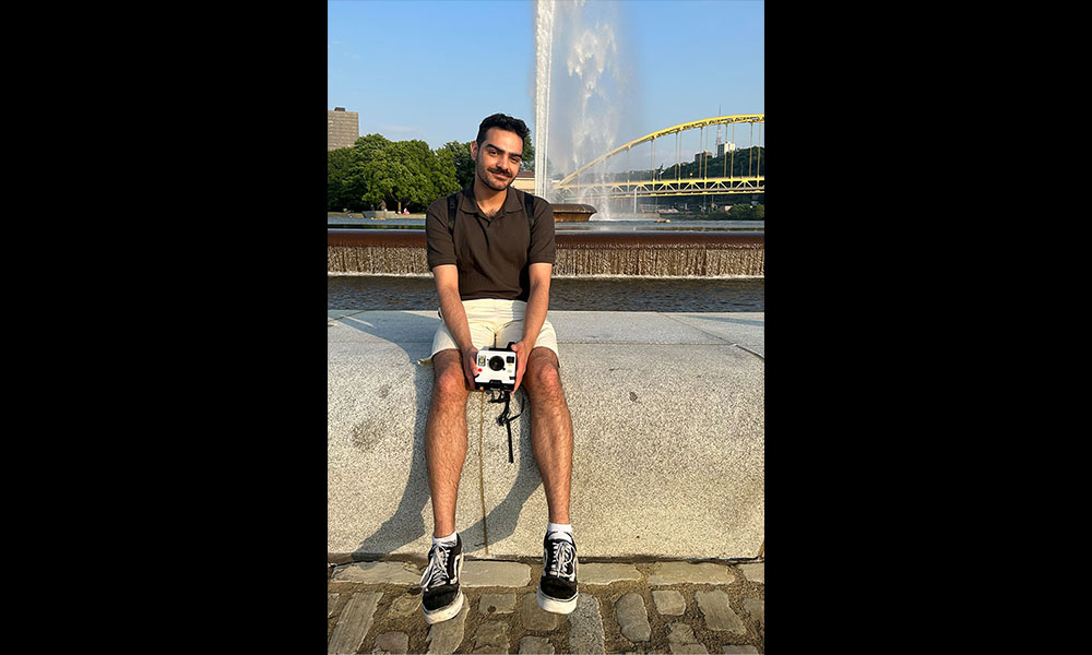 Japkaran Saroya sits on a concrete block in front of a yellow bridge during his placement in Pittsburgh