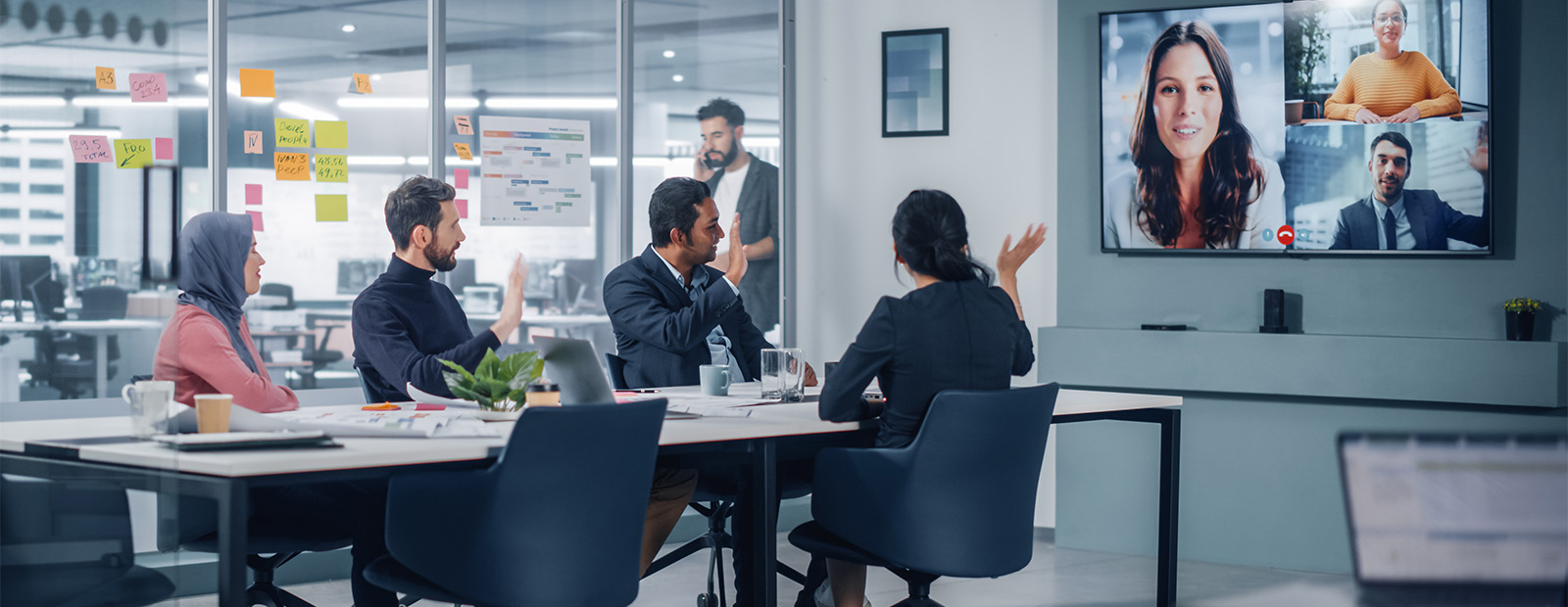 A group of colleagues sit around a boardroom table, but look at a wall-mounted monitor where remote workers are also involved in the meeting.