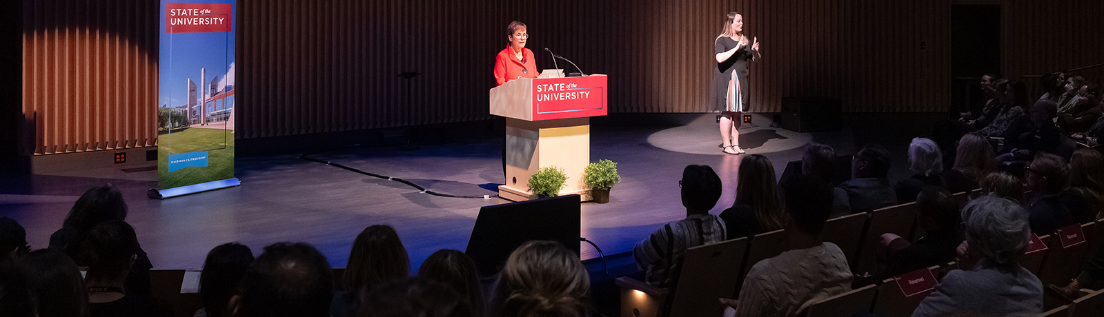 Dr. Annette Trimbee stands at the podium wearing a red jacket. A sign language interpreter is with her on stage.