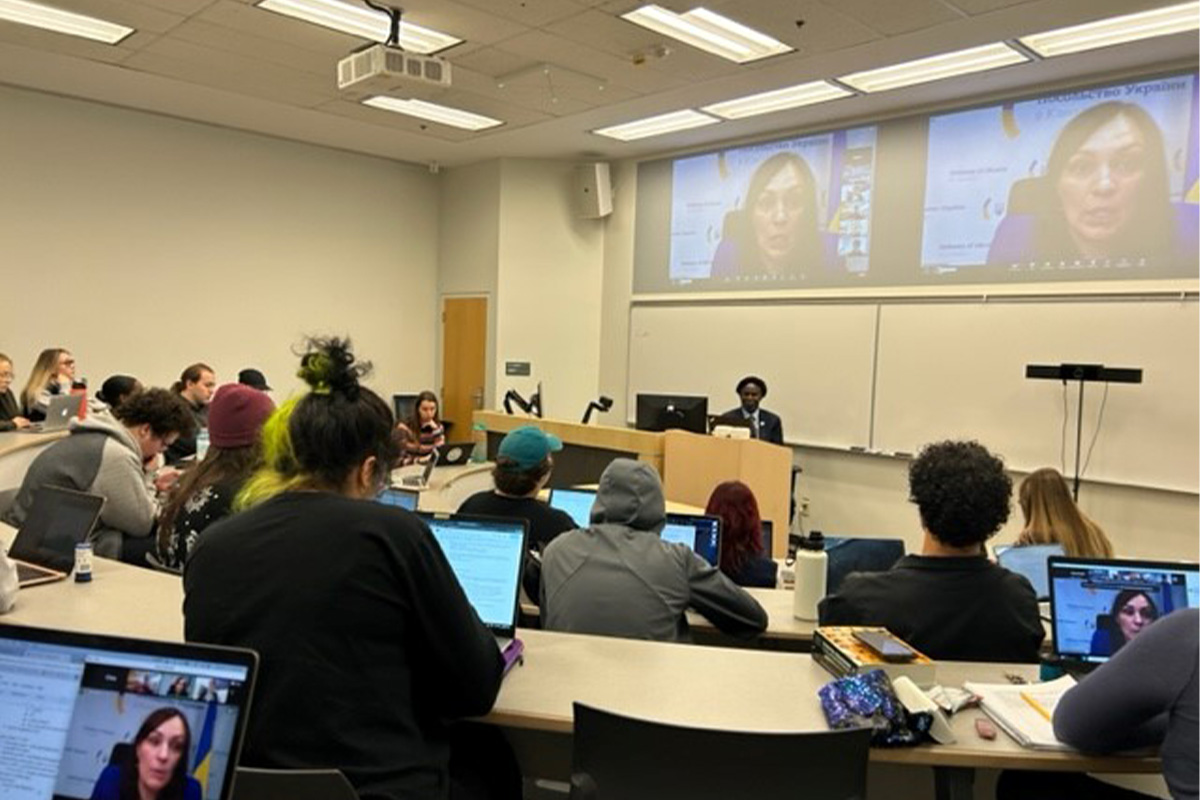 Students sit in a classroom and look toward a video chat window, where Her Excellency Yuliya Kovaliv, Ukraine Ambassador to Canada is speaking