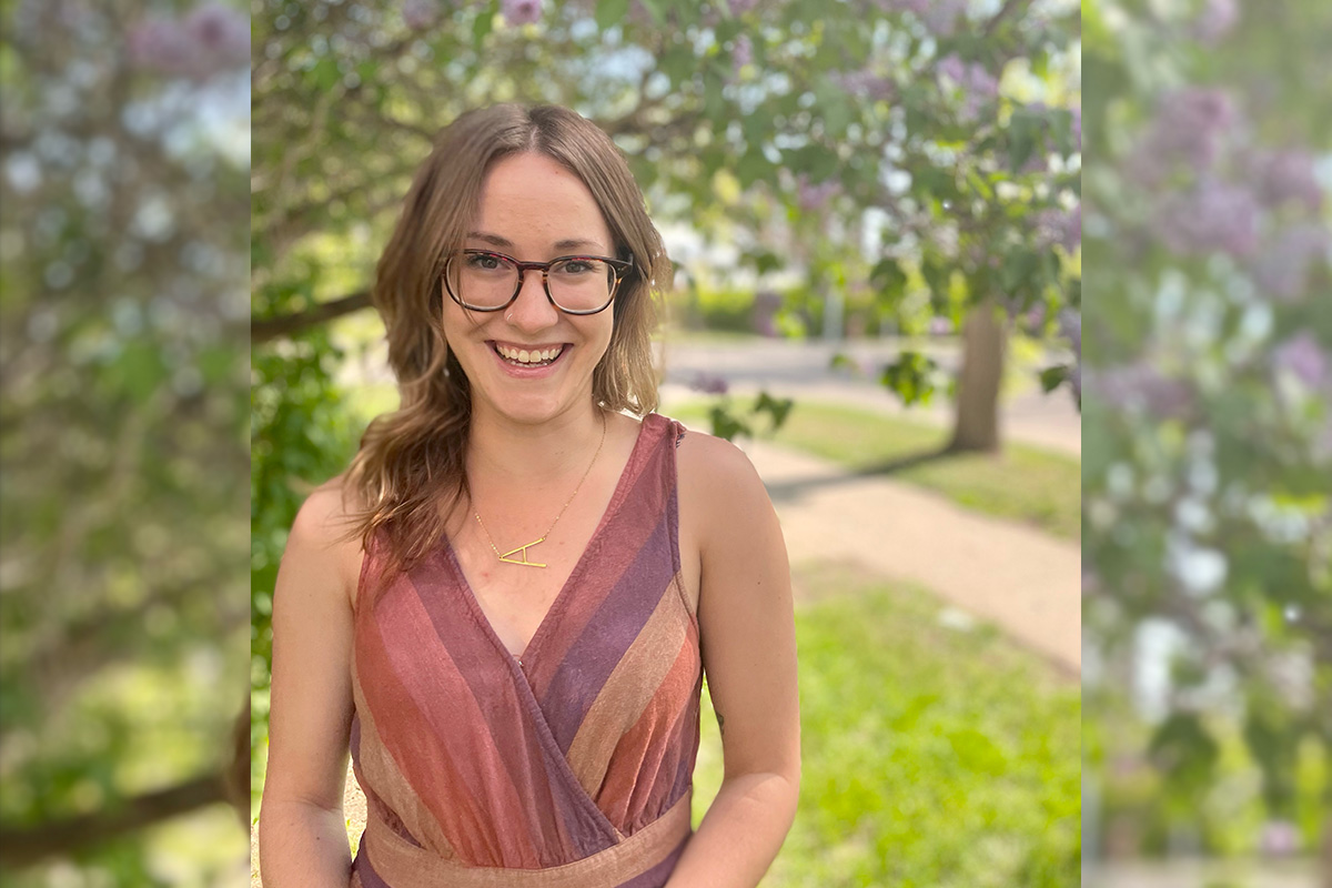 Alexandria wears a brown sleeveless shirt and stands in front of a green leafy tree.