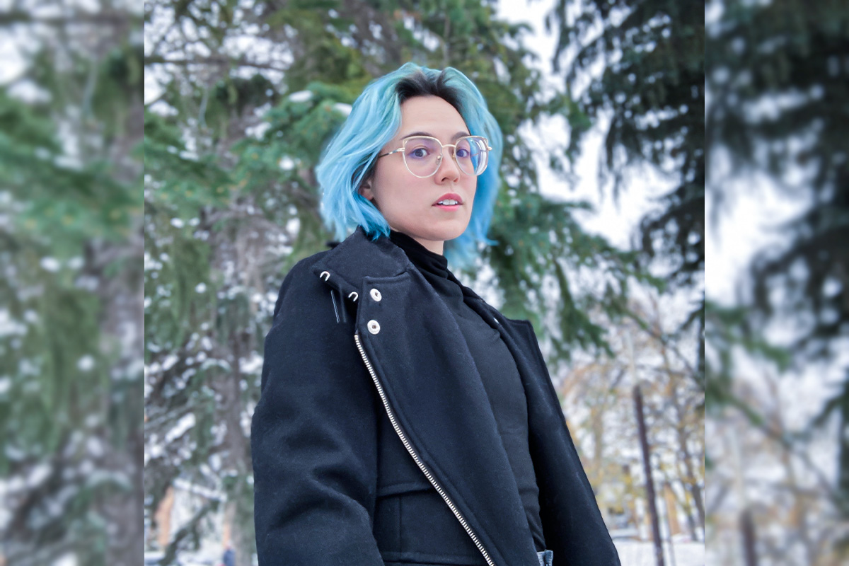 A woman with blue hair wearing a black leather jacket stands in front of snow-covered trees.