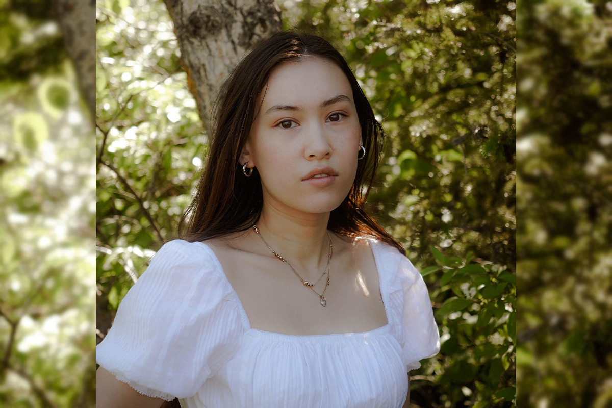 Bridgette wears a white shirt and looks at the camera with a serious expression, green trees behind her.