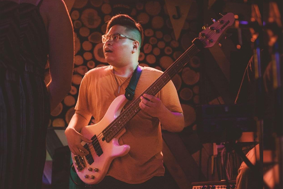 Jeremie wears a yellow tee shirt and plays a white bass guitar on stage.