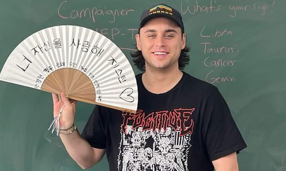 Justyn stands in front of a chalkboard holding a Korean fan