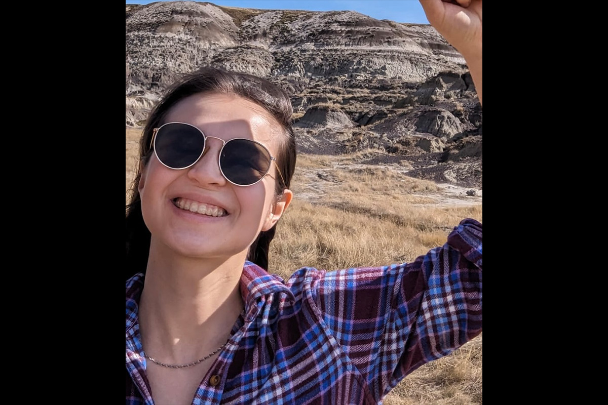 A woman with brown hair wears sunglasses and smiles, standing in front of badlands.