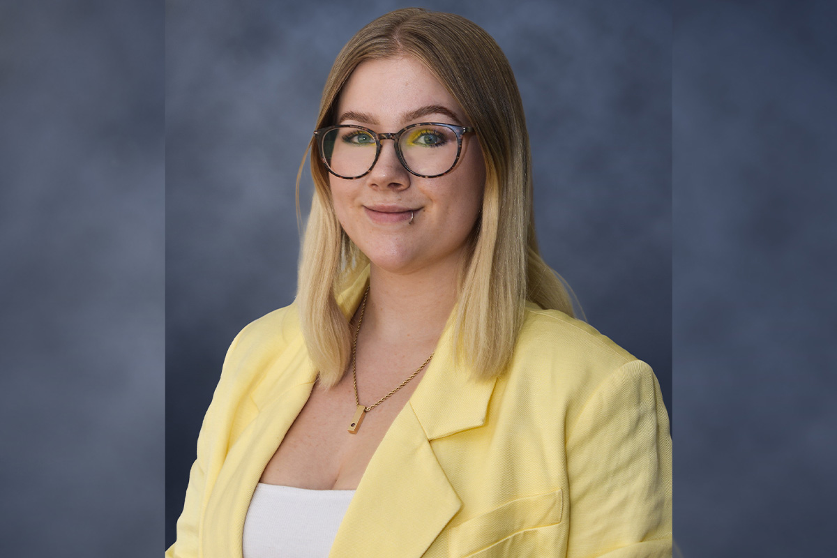 A blonde woman with black-framed glasses wears a yellow blazer in a professional headshot.