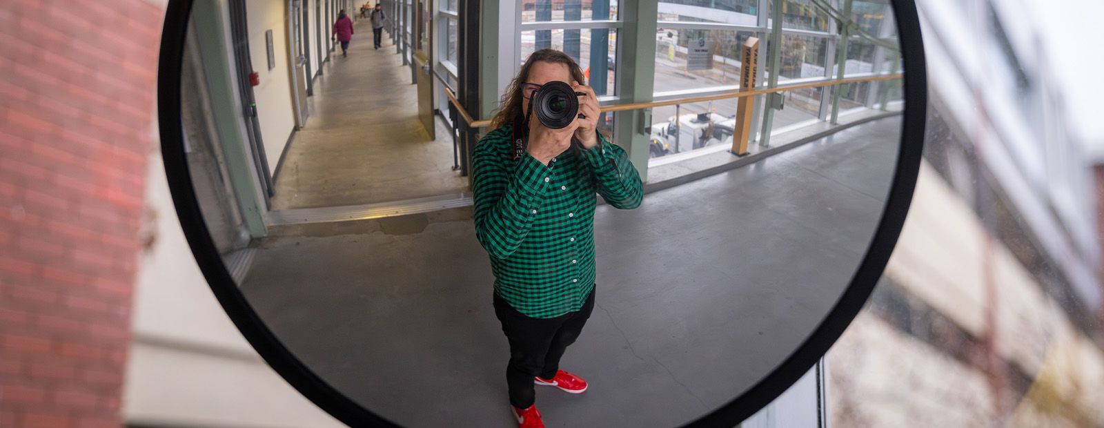 A self-portrait of Steven, his camera covering his face, taken in the reflection of a mirror in the corner of the 109 St. Pedway. He wears a green and black plaid shirt and jeans.