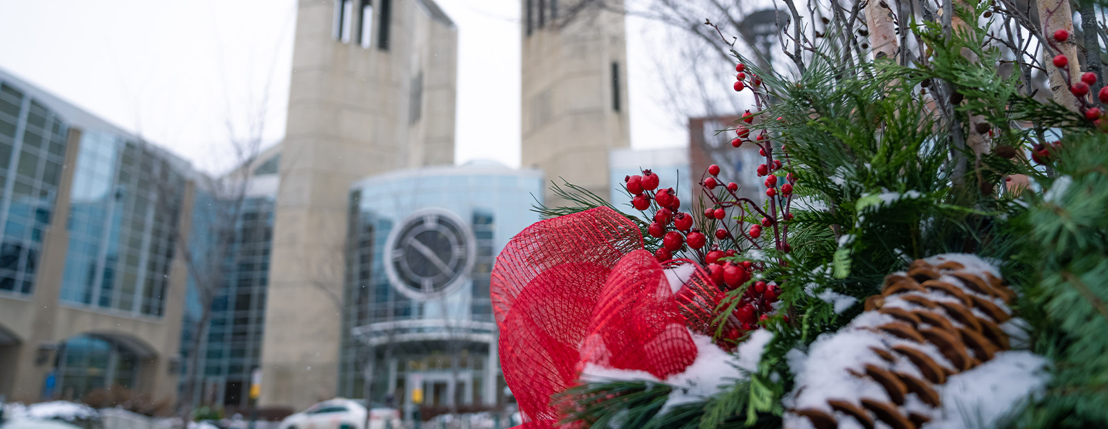 A holiday planter filled with red ribbons and pinecones is covered in snow, with the clocktower entrance to MacEwan in the background.