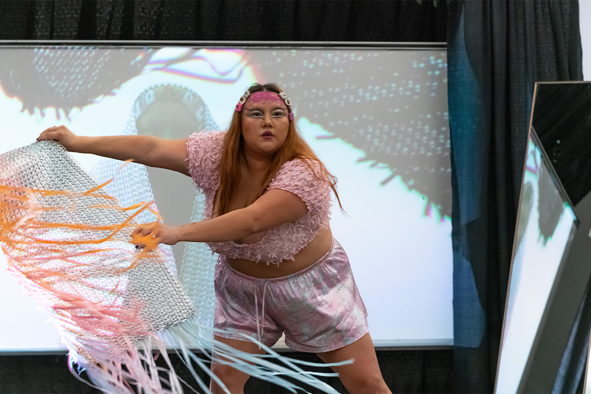 A woman with gemstones on her forehead and extravagant eyeliner wears a pink top and shorts, dancing on a stage in the Allard Hall atrium.