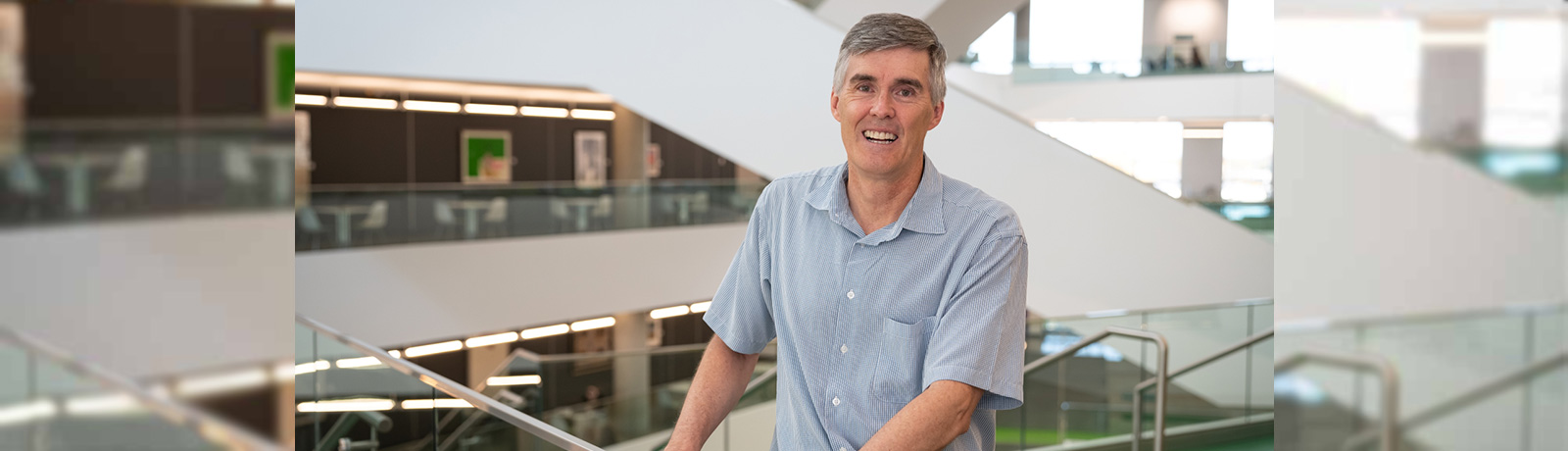 Dr. John Valentine wears a blue button-up tee shirt and leans against a glass railing in Allard Hall, smiling