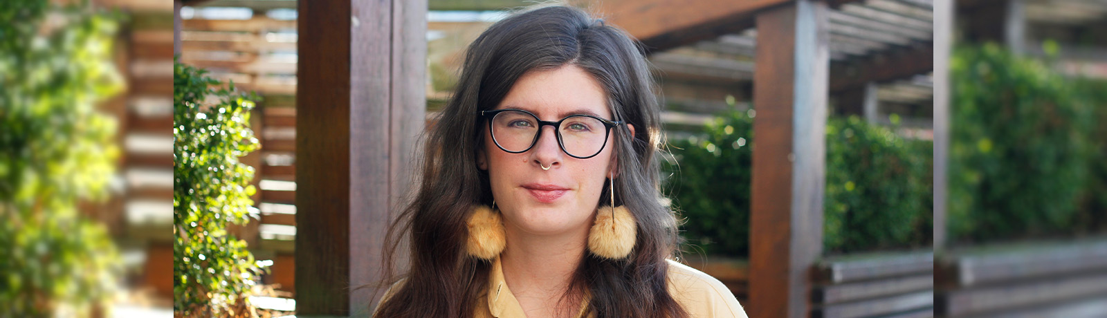 Jessica Johns wears a yellow shirt and yellow earrings, and stands in front of a wooden pergola.