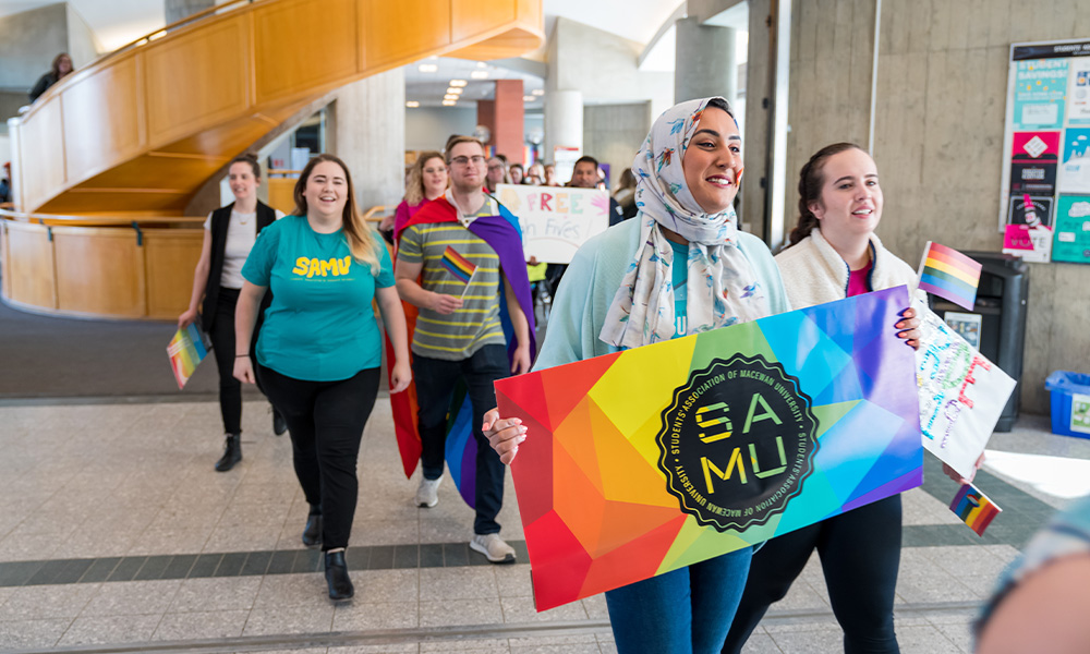 Students pass by the spiral staircase on campus, smiling and waving rainbow flags.