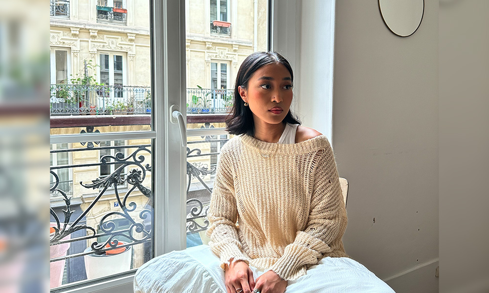 Abigail is sitting on the ledge of a window, indoors, that looks out onto other balconies. She looks away from the camera with her hands in her lap.