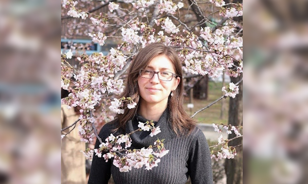 Ashley is wearing a grey sweater and standing within the branches of an apple tree in full bloom