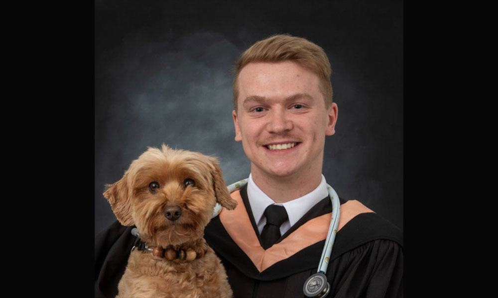 Dawson is standing in front of a grey background wearing his graduation gown and holding his dog 