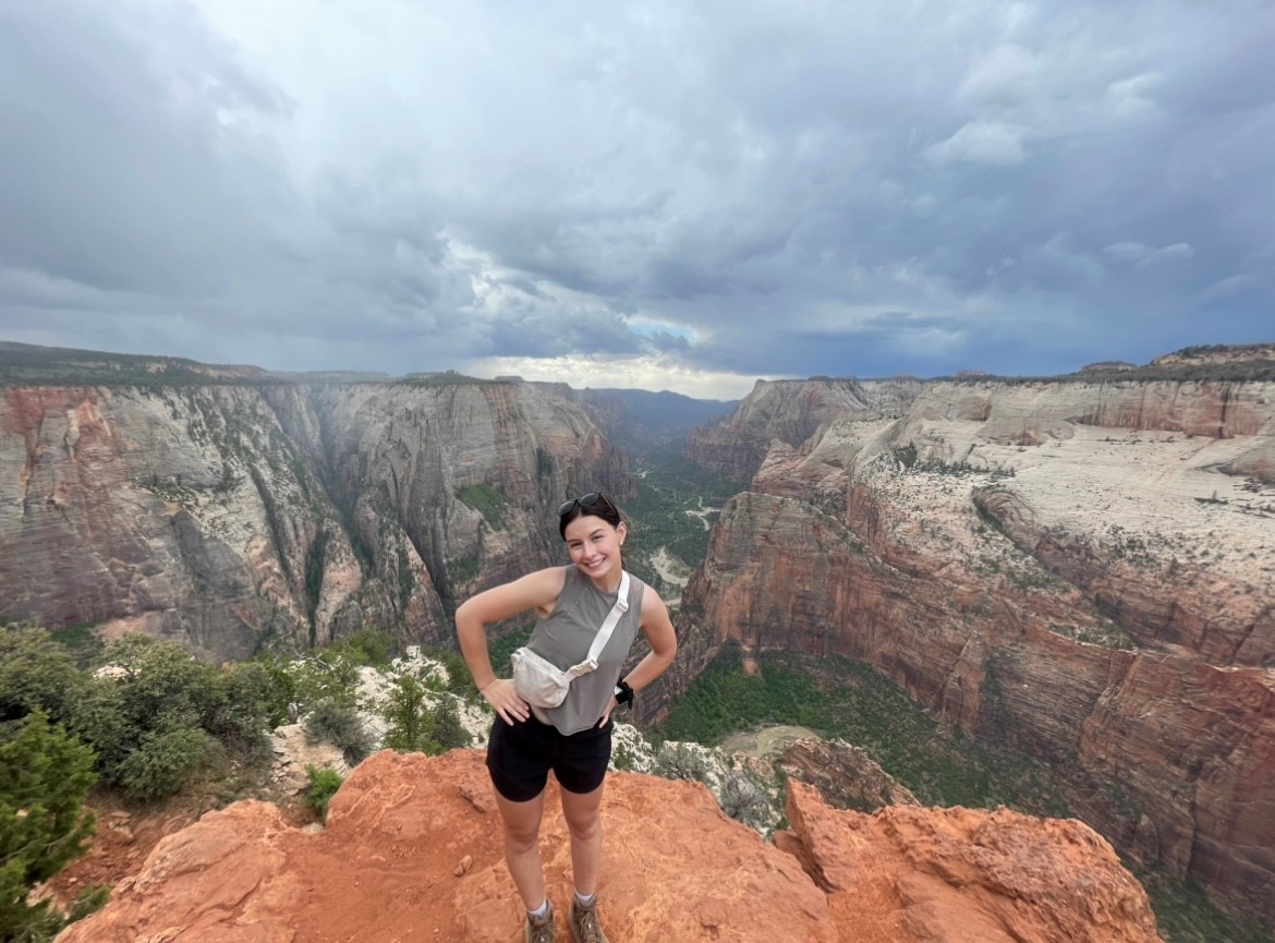 Madison is posing with hands on hips on a high cliff with a valley behind her